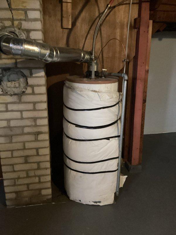 Conducted assessment for water heater replacement in 1940-era single-family home. Existing unit is 86 years old with visible deterioration, installed against brick wall in utility area with outdated insulation strapping and connection points. Inspection of plumbing components revealed aging pipe fittings integrated within wooden structural frame requiring modernization for proper installation compliance. Photos documented current installation configuration and structural considerations for replacement planning.