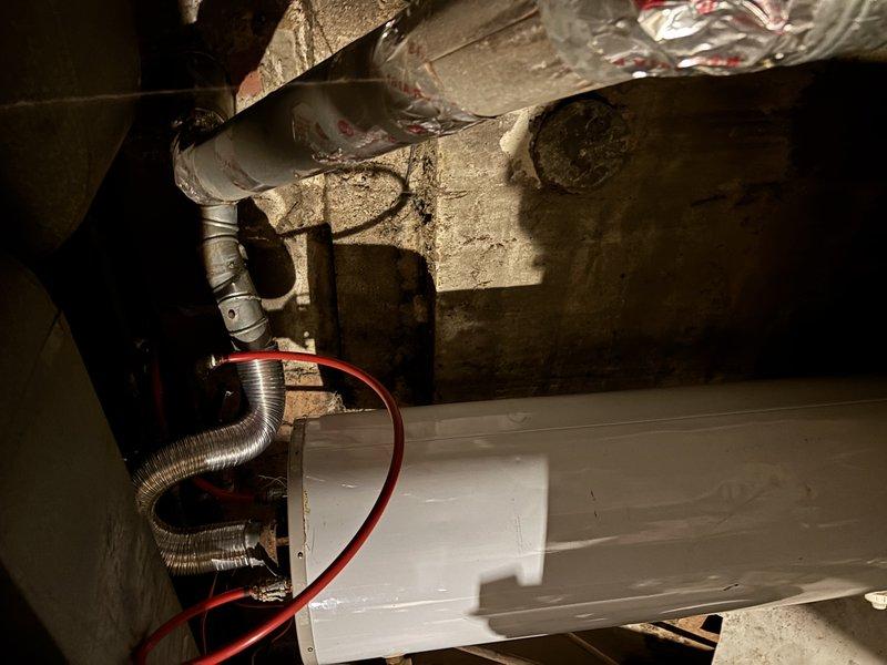Conducted water heater assessment for CWOW customer, documenting current installation and ventilation system. Existing unit is a white cylindrical water heater with copper piping and flexible metal vent pipe, installed against concrete block wall in basement utility area. Photos taken of complete mechanical space showing HVAC connections and water heater configuration for estimate preparation.