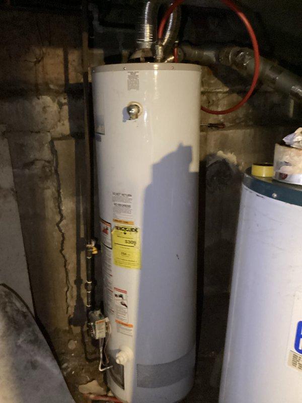 Conducted water heater assessment for CWOW customer, documenting current installation and ventilation system. Existing unit is a white cylindrical water heater with copper piping and flexible metal vent pipe, installed against concrete block wall in basement utility area. Photos taken of complete mechanical space showing HVAC connections and water heater configuration for estimate preparation.