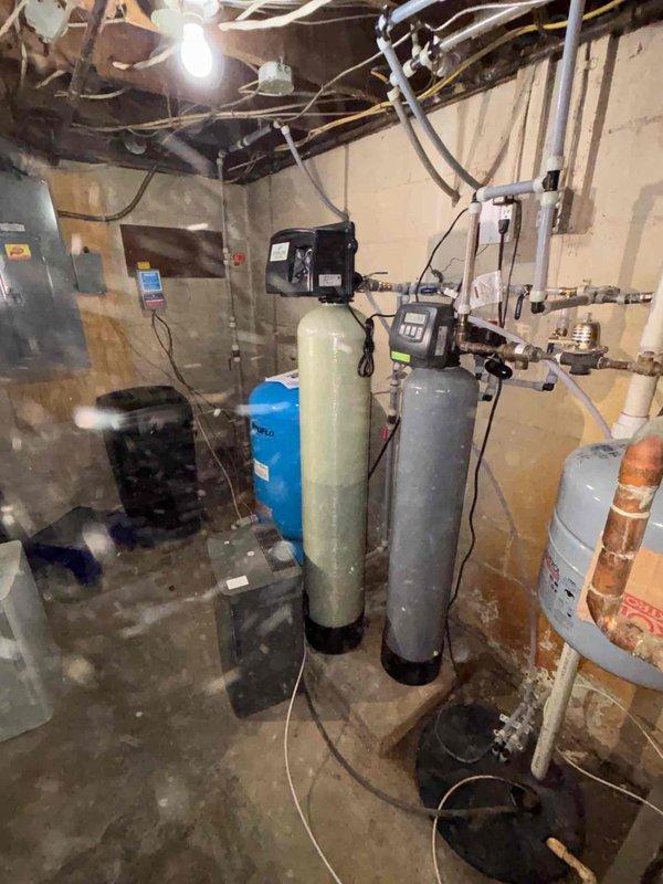 Completed installation of a comprehensive water treatment system in basement utility room, including multiple filtration tanks (one green, two gray with digital control panels) with all necessary copper and PVC piping connections. Additionally, installed sump pump system with basin embedded in concrete floor and proper discharge piping to prevent basement flooding.