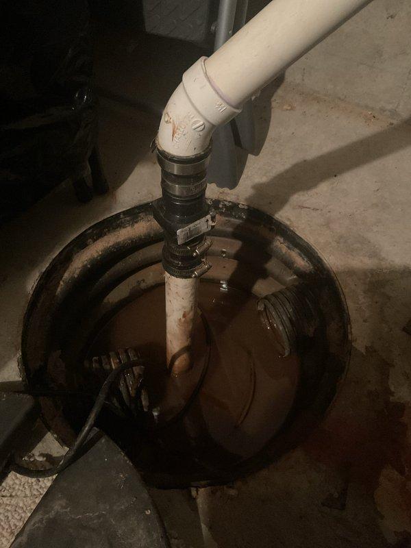 Conducted assessment of aging sump pump system in basement utility area. Inspected sump pit containing standing water with muddy appearance, examined PVC discharge pipe with black coupling/check valve, and evaluated submersible pump showing signs of age. Recommended replacement of existing system due to visible deterioration and potential for failure.