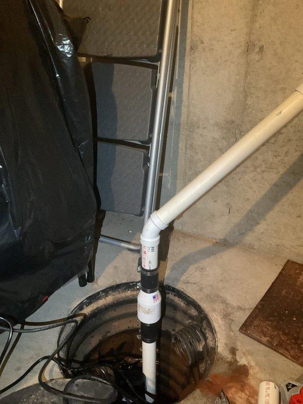 Conducted assessment of aging sump pump system in basement utility area. Inspected sump pit containing standing water with muddy appearance, examined PVC discharge pipe with black coupling/check valve, and evaluated submersible pump showing signs of age. Recommended replacement of existing system due to visible deterioration and potential for failure.