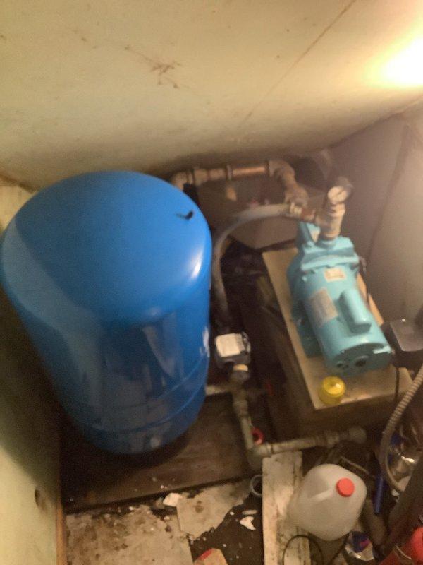Diagnosed non-functioning sump pump located in basement utility area. Inspection revealed faulty motor in the light blue pump unit mounted on wooden platform, with visible corrosion at copper pipe connections. Replaced pump motor assembly, tested system functionality, and verified proper operation before completing service.