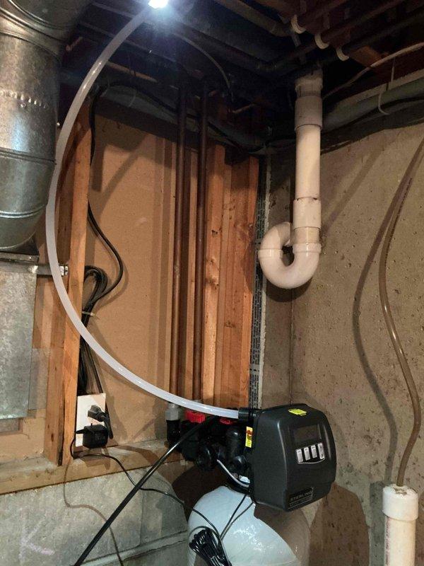 Successfully installed water softener system in basement utility area, connecting to existing plumbing infrastructure with proper PVC drain line configuration. System includes control unit with indicator lights and was tested for proper operation after installation.