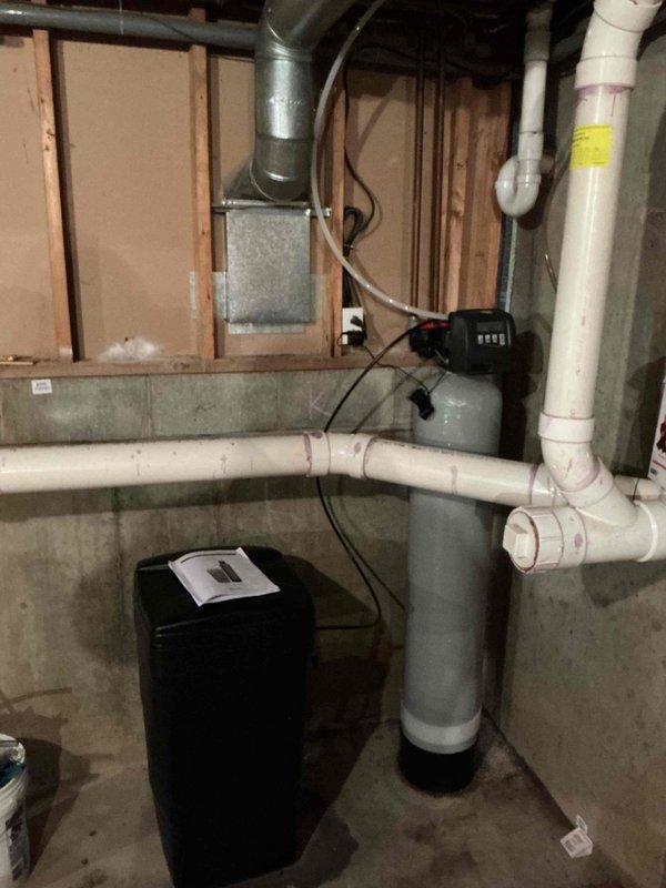Successfully installed water softener system in basement utility area, connecting to existing plumbing infrastructure with proper PVC drain line configuration. System includes control unit with indicator lights and was tested for proper operation after installation.