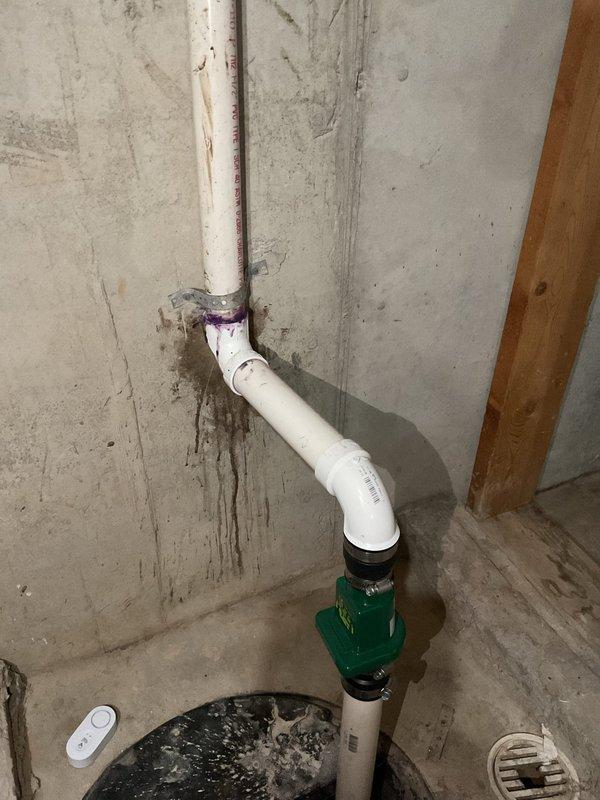 Performed estimate for backup sump pump installation in basement. Inspected existing sump pit containing standing water with visible float switch mechanism and documented current discharge plumbing configuration. Evaluated wall-mounted PVC discharge pipe system with installed check valve and noted signs of previous moisture issues requiring attention during potential installation.