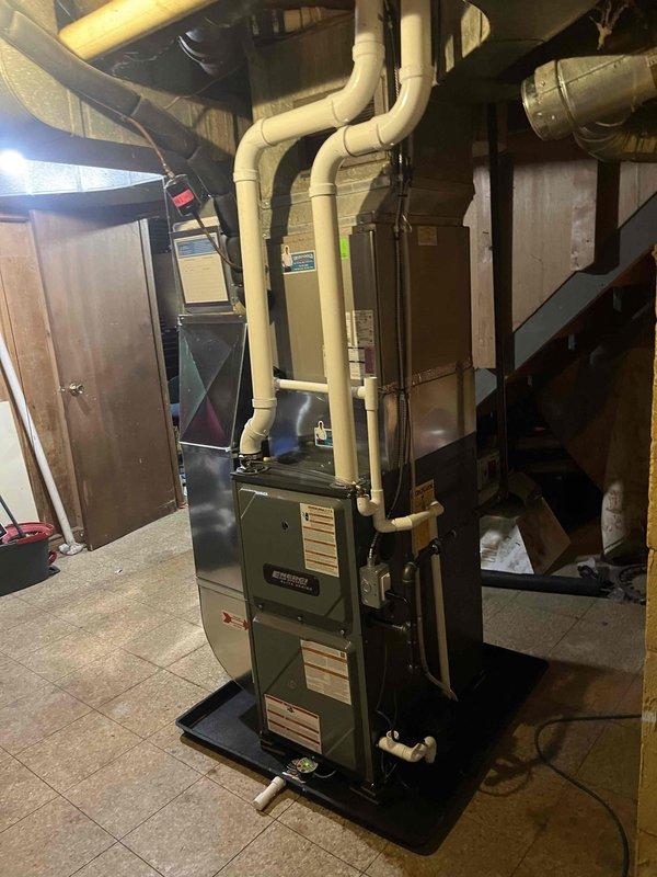 Completed installation of a new residential furnace in basement utility area. System includes furnace unit with PVC intake/exhaust venting, properly connected to existing ductwork with angular metal transitions. Unit was positioned on drip pan against concrete block wall with all electrical components and control systems connected according to manufacturer specifications.