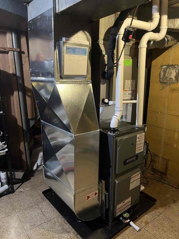 Completed installation of a new residential furnace in basement utility area. System includes furnace unit with PVC intake/exhaust venting, properly connected to existing ductwork with angular metal transitions. Unit was positioned on drip pan against concrete block wall with all electrical components and control systems connected according to manufacturer specifications.