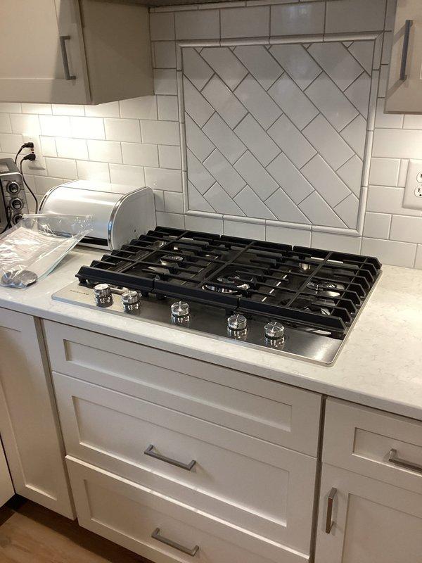 Conducted site assessment for gas cooktop installation and tankless water heater estimate. Inspected existing five-burner stainless steel cooktop integrated into white countertop with herringbone tile backsplash. Documented current kitchen configuration and appliance placement for accurate project planning and material requirements.