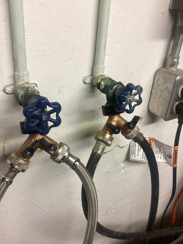 Responded to a plumbing leak service call for washing machine connections. Inspection revealed mineral buildup and corrosion at the connection points between PVC supply pipes and brass shutoff valves with blue handles. Replaced deteriorated braided supply lines, cleaned connection points, and verified proper operation with no further leakage.