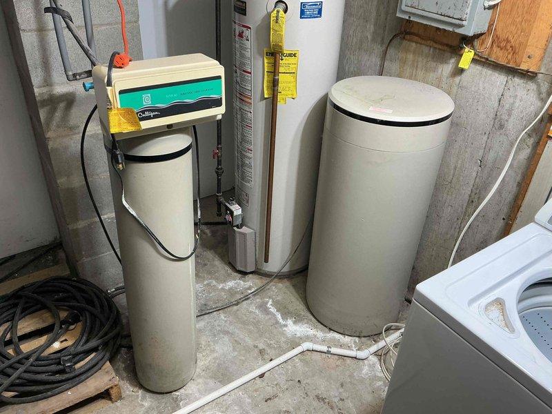 Conducted comprehensive assessment of client's water systems for replacement of water heater, water softener, and well pressure tank. Documented existing setup in utility area showing two cylindrical treatment tanks with control systems, current water heater installation, and associated plumbing connections. Also noted potential water leak at toilet supply line connection with visible moisture staining at base, which will require attention during system upgrades.
