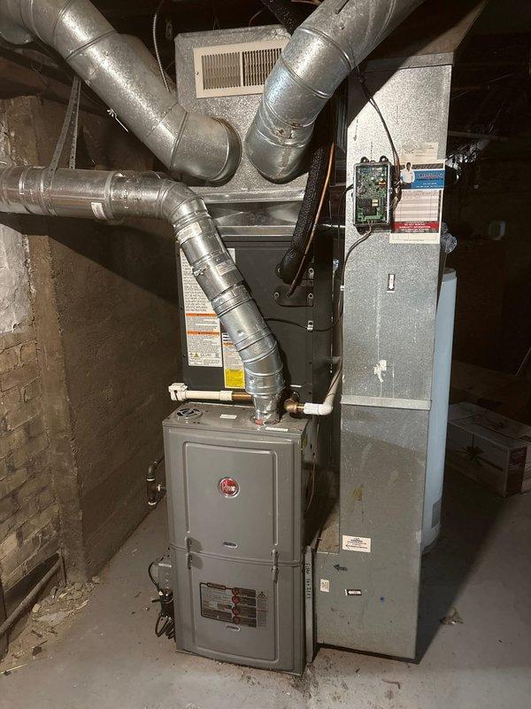 Successfully completed installation of new Amana outdoor air conditioning unit, properly positioned on concrete pad with copper refrigerant lines connected to the home. System includes Rheem furnace and air handler with appropriate galvanized metal ductwork installed in utility area, all components verified operational with proper connections.