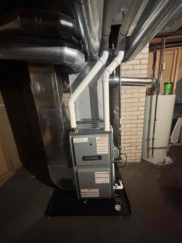 Successfully completed installation of new gray furnace unit in basement utility room, including proper connection of metal ductwork for air distribution and installation of PVC condensate drain lines. System was mounted on appropriate black platform with all warning labels properly affixed, and installation includes complete integration with existing ductwork configuration.