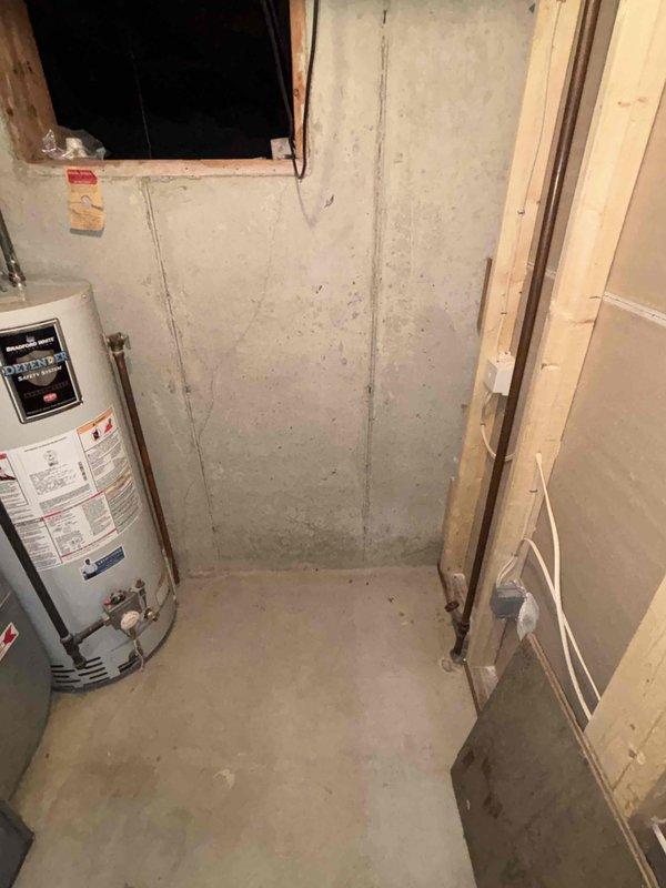 Completed water heater estimate for a 20-year-old Bradford White unit located in an unfinished utility area with concrete walls and floor. Documented current installation configuration including existing plumbing connections and surrounding space constraints for potential replacement options.
