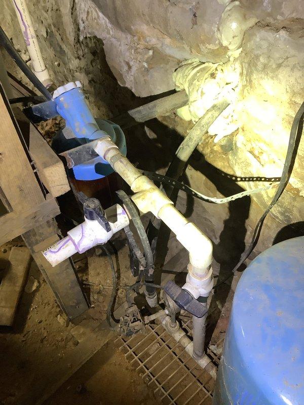 Inspected water system in vacant single-family residence and documented extensive issues with the well water system. Found bladder tank with visible perforation requiring complete replacement, along with a malfunctioning toilet and leaking valve. Photographic documentation captured the existing PVC pipe network, connection points, and the damaged blue pressure tank showing significant wear and corrosion at its base.