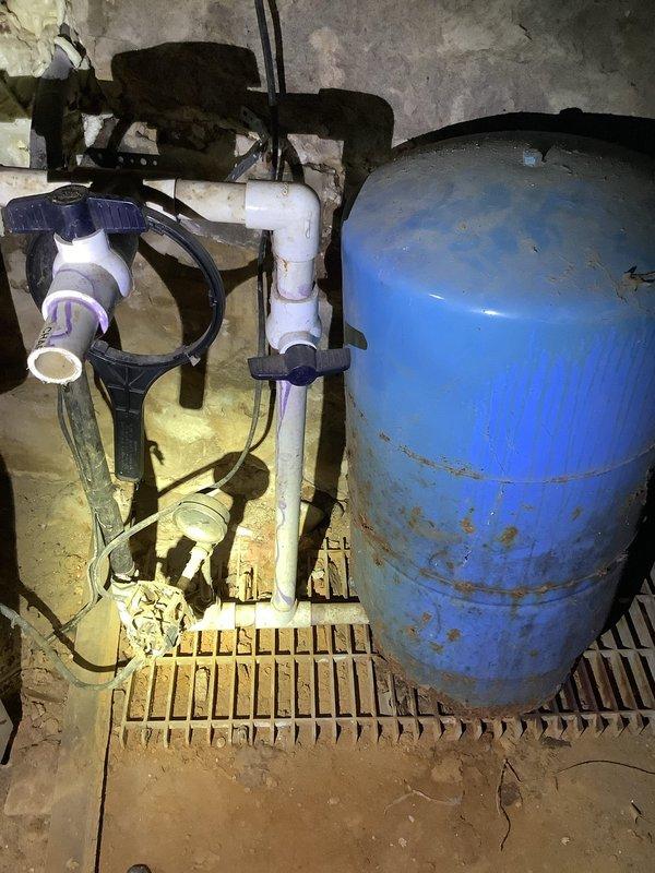 Inspected water system in vacant single-family residence and documented extensive issues with the well water system. Found bladder tank with visible perforation requiring complete replacement, along with a malfunctioning toilet and leaking valve. Photographic documentation captured the existing PVC pipe network, connection points, and the damaged blue pressure tank showing significant wear and corrosion at its base.