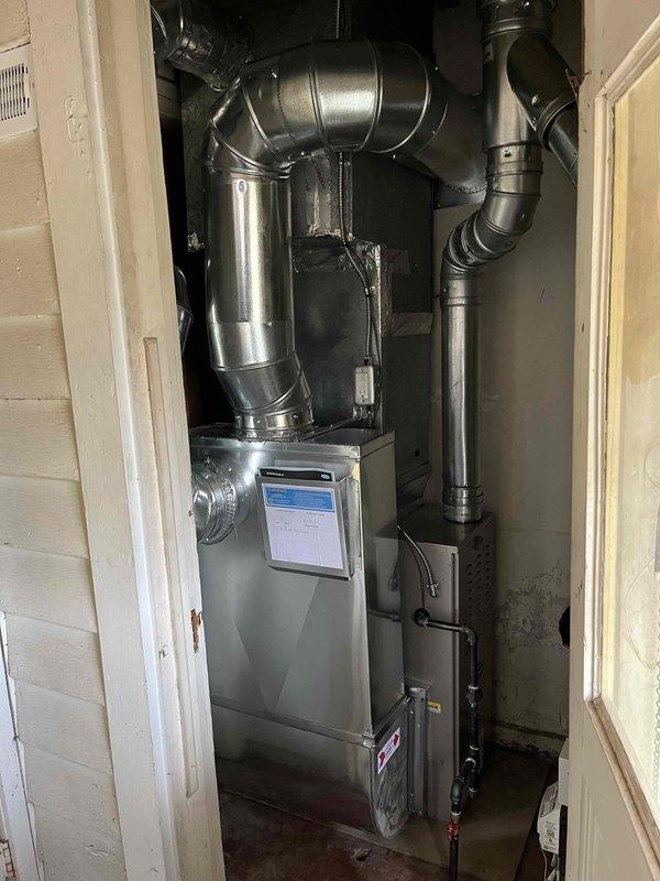 Successfully completed installation of new furnace unit with associated ductwork in utility area. System features digital control interface and proper connections for all components with secure metal tape seals at junction points. Installation verified complete and operational.