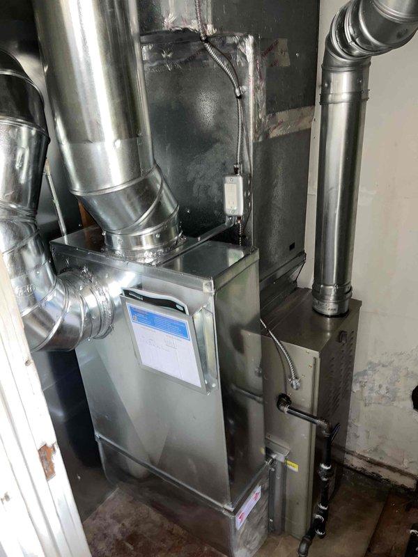 Successfully completed installation of new furnace unit with associated ductwork in utility area. System features digital control interface and proper connections for all components with secure metal tape seals at junction points. Installation verified complete and operational.