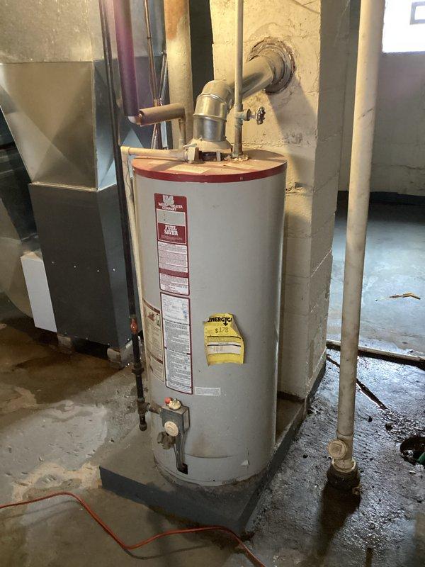 Performed comprehensive assessment of existing water heater system in basement utility room. Unit shows signs of age with corrosion at connection points, mineral buildup, and water damage/staining on adjacent wall. Documented current installation including vent pipe, pressure relief valves, and supply connections to provide appropriate replacement options.