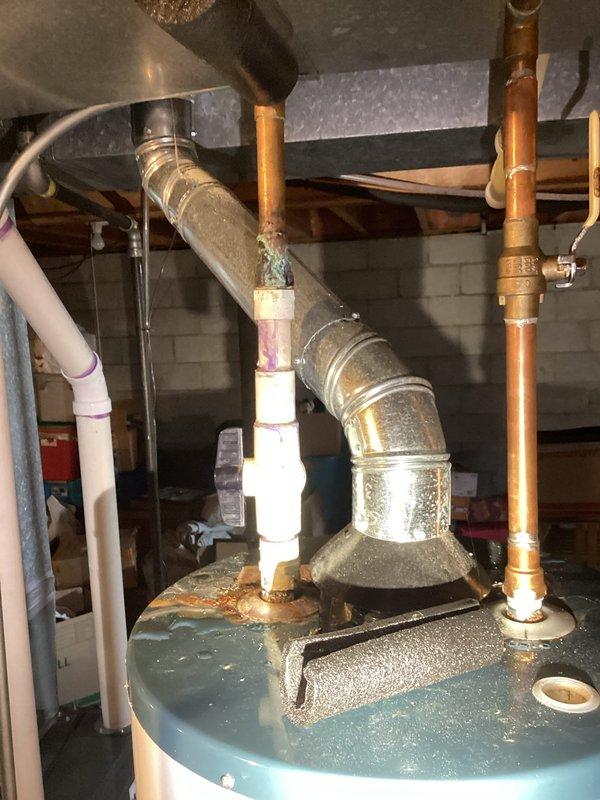 # Plumbing Leak Inspection Report

Responded to service call for leaking pipe investigation. Upon arrival, conducted thorough visual inspection of water heater and associated plumbing connections in utility area. Inspection revealed significant rust and corrosion on water heater top surface, particularly concentrated around pipe connection points. Multiple copper supply lines and galvanized fittings showed signs of age-related deterioration with visible patina and surface oxidation.

Primary concern identified at base of white PVC discharge pipe penetrating through blue metal drip pan. Extensive rust and corrosion present at pipe-to-pan interface, with brown and orange discoloration spreading across metal surface. Water staining and moisture damage visible on surrounding areas, indicating active or recurring leak condition. Rust buildup and textured corrosion patterns suggest prolonged water exposure requiring immediate attention.

Documented conditions with photographs showing water h