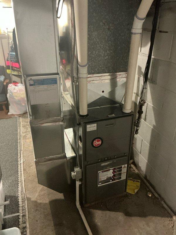 Conducted on-site assessment of existing 52-year-old Luxaire residential HVAC system for complete air conditioning replacement estimate. Evaluated basement-installed furnace and air handler configuration, documenting current gray metal furnace unit with upward-routed white PVC condensate drainage and cylindrical ductwork connections. Inspected outdoor condenser unit mounted on mulch bed against brick exterior wall, noting significant weathering, discoloration, and wear on metal casing consistent with age of system. Photographed both indoor and outdoor equipment to document current installation conditions, equipment specifications, and routing of electrical conduit and drainage lines for replacement planning purposes.