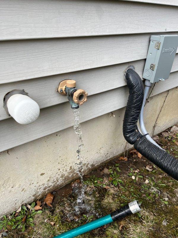 Responded to service call for outdoor faucet with suspected freeze damage after hose was left connected throughout winter season. Upon arrival, inspected dual hose bib installation on exterior wall with vinyl siding. Found one faucet equipped with foam insulation cover while second unit remained exposed. Conducted operational test which revealed active water leak from affected hose bib, confirming freeze damage to internal valve components. 

Documented pre-existing conditions showing water streaming from spout and splashing onto concrete foundation, indicating compromised valve seat or stem assembly due to ice expansion. Verified both faucets for accessibility and assessed extent of damage. One unit showed proper winterization with protective cover in place, while damaged unit exhibited typical failure pattern consistent with water remaining in valve body during freezing temperatures.

Photographed damaged faucet during active leak to document failure mode and surrounding plumbing inf