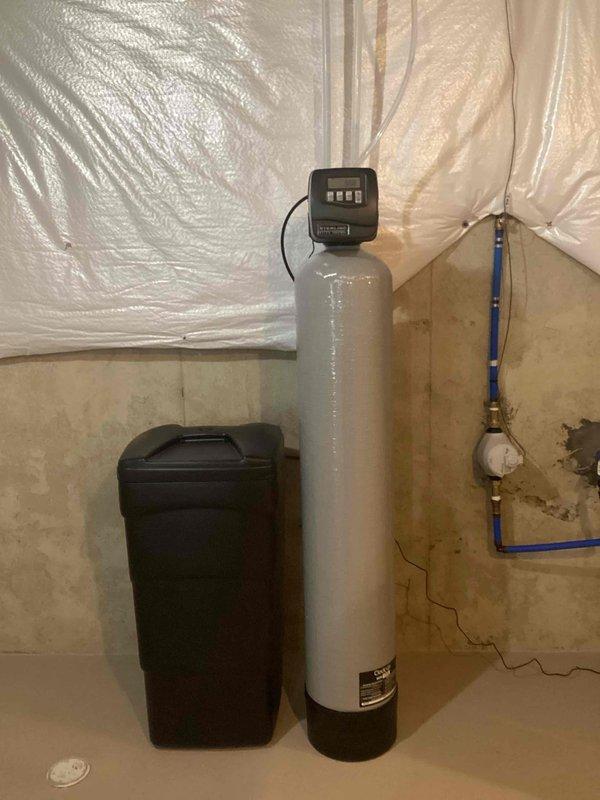 Completed installation of residential water softener system in basement utility area. Installed main white cylindrical softener tank with digital control head and black brine tank for salt storage, positioning equipment against foundation wall with proper drainage access. Ran blue PEX water supply lines from basement through attic space to integrate system with existing plumbing infrastructure, securing all piping with appropriate clips and brackets. Connected system to existing copper/brass valve assembly and installed spigot as specified. All components tested and operational upon completion.