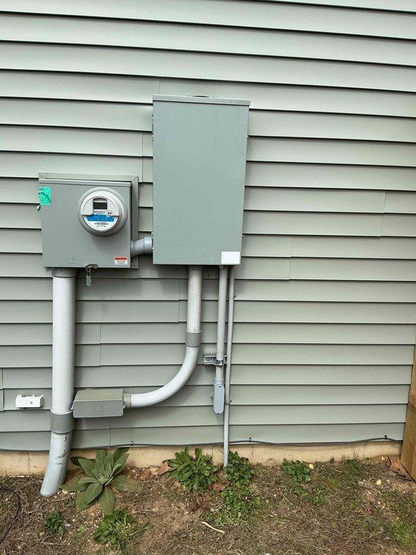Completed installation of electrical interlock kit on residential service panel. Installed interlock device between existing meter base and electrical disconnect panel, connecting components with white PVC conduit and appropriate mounting hardware secured to vinyl siding. System now allows for safe generator connection with proper load transfer functionality.