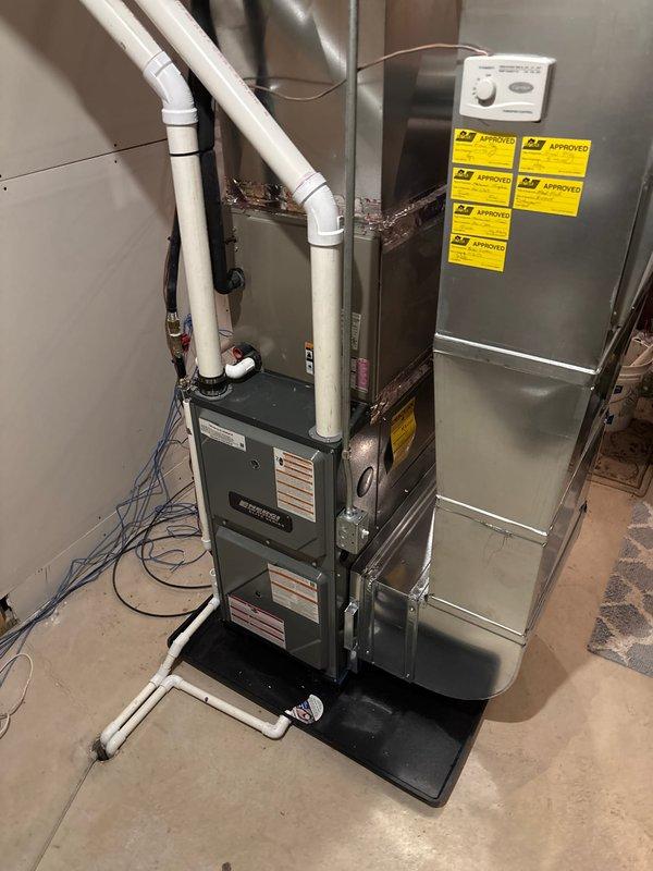 Completed full furnace and air handler installation in basement utility area. Installed new Carrier HVAC system including dark gray furnace unit with white PVC condensate drain and venting lines routed to ceiling, paired with tall silver metallic air handler mounted on black protective platform. Connected all refrigerant lines, electrical conduits, and ductwork; installed beige humidifier attachment to top of air handler with proper ventilation and indicator light. Verified all system connections, tested operation, and ensured proper drainage and venting configuration per manufacturer specifications.
