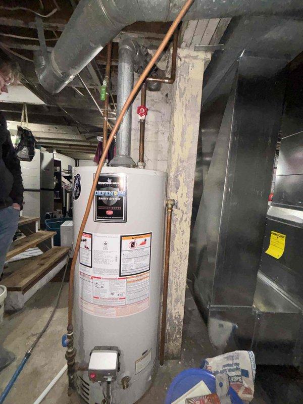 Responded to complaint of popping and banging noises from Bradford water heater (circa 2011). Upon inspection, unit was still producing hot water but exhibited signs of sediment accumulation causing the reported sounds. Documented existing plumbing configuration including copper supply lines, shutoff valves, and HVAC ductwork in utility area. Photographed water heater installation and surrounding mechanical systems for service records. Unit remains operational with continued hot water production despite acoustic issues typical of mineral buildup in aging tank.