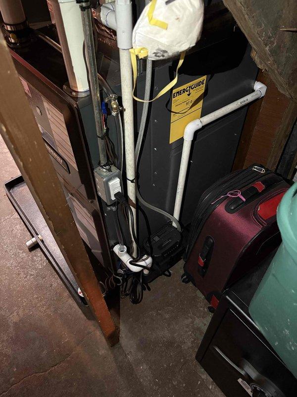 Completed full furnace installation in basement utility area. Installed new black air handler unit with proper electrical connections and bundled wiring. Routed white PVC condensate drainage and venting lines vertically along equipment. Connected silver metallic ductwork with appropriate elbows and straight sections to distribution system. Mounted condensate pump at base of air handler unit. Positioned equipment on protective black rubber mat over concrete floor. Installed gray metal auxiliary component adjacent to main furnace unit. Mounted Honeywell control plate on wall for system operation. Verified all manufacturer specification labels properly affixed to equipment. Ensured proper clearances and access for future maintenance. System tested and operational upon completion.