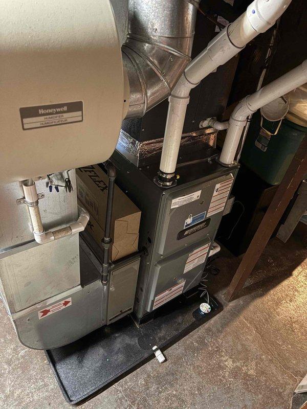 Completed full furnace installation in basement utility area. Installed new black air handler unit with proper electrical connections and bundled wiring. Routed white PVC condensate drainage and venting lines vertically along equipment. Connected silver metallic ductwork with appropriate elbows and straight sections to distribution system. Mounted condensate pump at base of air handler unit. Positioned equipment on protective black rubber mat over concrete floor. Installed gray metal auxiliary component adjacent to main furnace unit. Mounted Honeywell control plate on wall for system operation. Verified all manufacturer specification labels properly affixed to equipment. Ensured proper clearances and access for future maintenance. System tested and operational upon completion.