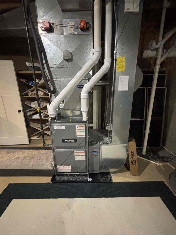 Completed full furnace installation in basement utility area. Installed new gray metal furnace unit on protective mat with proper placement and leveling. Connected and secured metal ductwork system to unit with appropriate fittings and orange connection points. Installed white PVC venting pipes from top of unit with proper elbows and directional routing for exhaust and intake. Verified all manufacturer labels and warning stickers properly affixed to unit panels. Ensured electrical connections, safety switches, and controls properly installed and functional. System tested and confirmed operational upon completion.