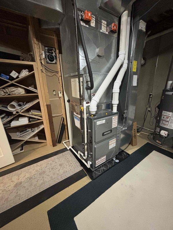 Completed full furnace installation in basement utility area. Installed new gray metal furnace unit on protective mat with proper placement and leveling. Connected and secured metal ductwork system to unit with appropriate fittings and orange connection points. Installed white PVC venting pipes from top of unit with proper elbows and directional routing for exhaust and intake. Verified all manufacturer labels and warning stickers properly affixed to unit panels. Ensured electrical connections, safety switches, and controls properly installed and functional. System tested and confirmed operational upon completion.