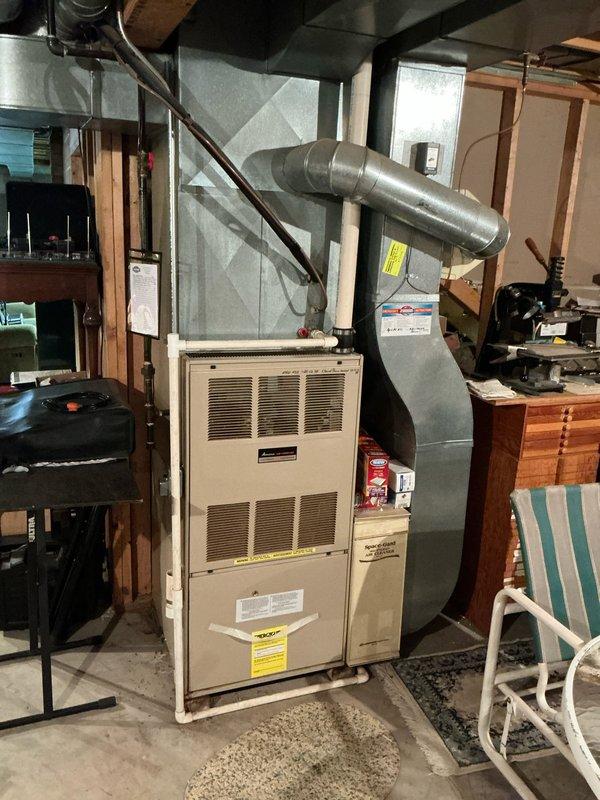 Completed on-site evaluation for full HVAC system replacement including furnace and air conditioning unit. Assessed existing beige residential furnace located in basement utility area with connected ductwork, gas lines, and copper refrigerant tubing. Evaluated outdoor AC condenser unit positioned in under-deck crawl space area with wire mesh protective grille, mounted on gravel base against painted concrete block wall. Documented current system configuration, electrical connections, ductwork routing, and installation conditions. Photographed both interior and exterior equipment to support replacement proposal and equipment specifications.