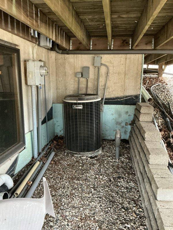 Completed on-site evaluation for full HVAC system replacement including furnace and air conditioning unit. Assessed existing beige residential furnace located in basement utility area with connected ductwork, gas lines, and copper refrigerant tubing. Evaluated outdoor AC condenser unit positioned in under-deck crawl space area with wire mesh protective grille, mounted on gravel base against painted concrete block wall. Documented current system configuration, electrical connections, ductwork routing, and installation conditions. Photographed both interior and exterior equipment to support replacement proposal and equipment specifications.