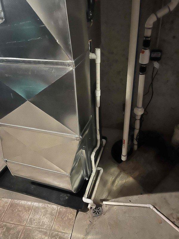 Completed full furnace installation in basement utility area, including mounting of new air handler unit with custom angular sheet metal ductwork configuration. Installed white PVC condensate drainage piping and exhaust/intake venting system running vertically through ceiling joists, connected metal supply ductwork to distribution system, and positioned unit on drip pan above existing floor drain. All equipment properly secured, labeled, and tested for operation with professional finish work completed.