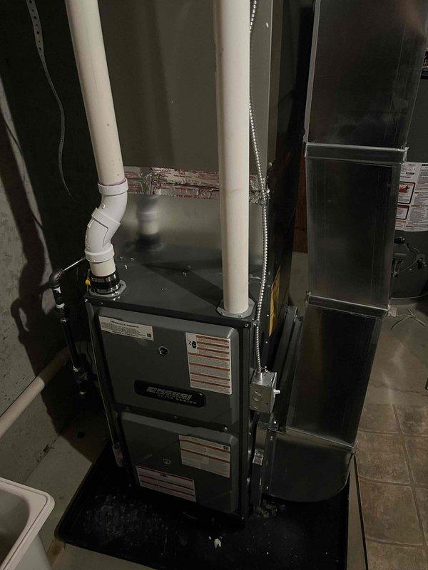 Completed full furnace installation in basement utility area, including mounting of new air handler unit with custom angular sheet metal ductwork configuration. Installed white PVC condensate drainage piping and exhaust/intake venting system running vertically through ceiling joists, connected metal supply ductwork to distribution system, and positioned unit on drip pan above existing floor drain. All equipment properly secured, labeled, and tested for operation with professional finish work completed.