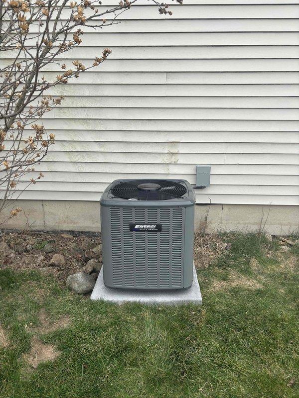 Completed full HVAC system replacement including outdoor condensing unit and indoor air handler. Removed existing equipment and installed new gray condensing unit on concrete pad against exterior wall with proper clearances maintained. Mounted unit features top-discharge fan configuration with horizontal louver design for optimal airflow. Ran electrical conduit and wiring from exterior disconnect box to new condenser, ensuring code-compliant installation. 

Inside mechanical room, installed new vertical furnace unit with integrated air handler, connecting to existing ductwork system using metallic supply plenum and trunk lines. Established refrigerant line set between outdoor and indoor units with proper insulation and securing. Connected condensate drain lines using PVC piping routed to approved drainage point. Verified all electrical connections at furnace unit and tested voltage at disconnect. Performed system startup procedures including refrigerant charge verification, airflow mea