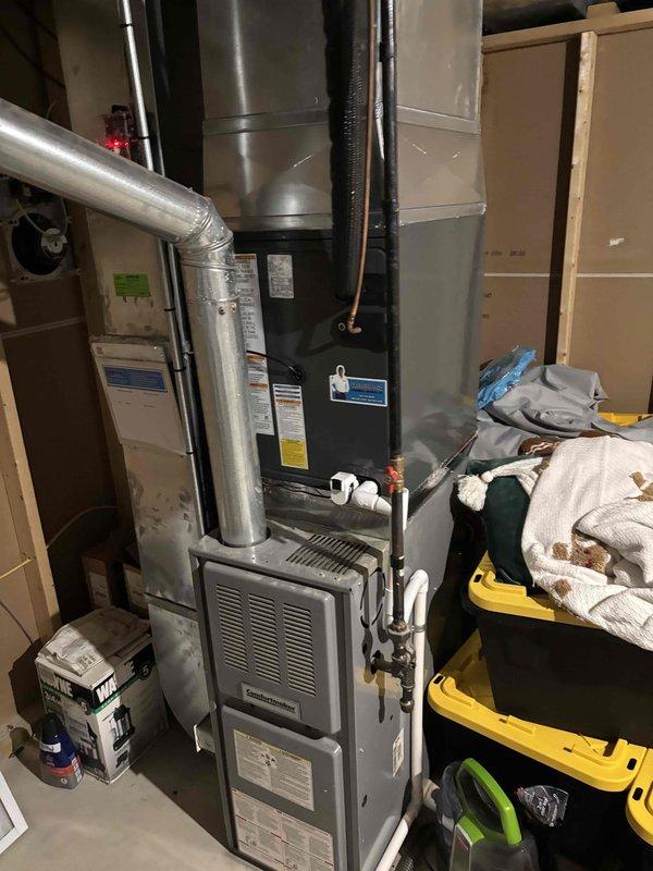 Completed full HVAC system replacement including outdoor condensing unit and indoor air handler. Removed existing equipment and installed new gray condensing unit on concrete pad against exterior wall with proper clearances maintained. Mounted unit features top-discharge fan configuration with horizontal louver design for optimal airflow. Ran electrical conduit and wiring from exterior disconnect box to new condenser, ensuring code-compliant installation. 

Inside mechanical room, installed new vertical furnace unit with integrated air handler, connecting to existing ductwork system using metallic supply plenum and trunk lines. Established refrigerant line set between outdoor and indoor units with proper insulation and securing. Connected condensate drain lines using PVC piping routed to approved drainage point. Verified all electrical connections at furnace unit and tested voltage at disconnect. Performed system startup procedures including refrigerant charge verification, airflow mea