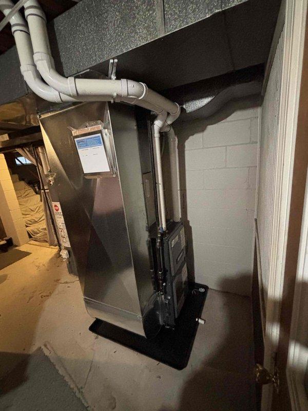 Completed full furnace replacement in basement mechanical room. Installed new Carrier HVAC unit with proper venting configuration using white PVC piping routed horizontally along upper wall section. Positioned condensate drain pan at base of unit on concrete floor. Verified all ductwork connections to existing black ductwork system and ensured proper clearances from adjacent water heater. System tested and operational upon completion.