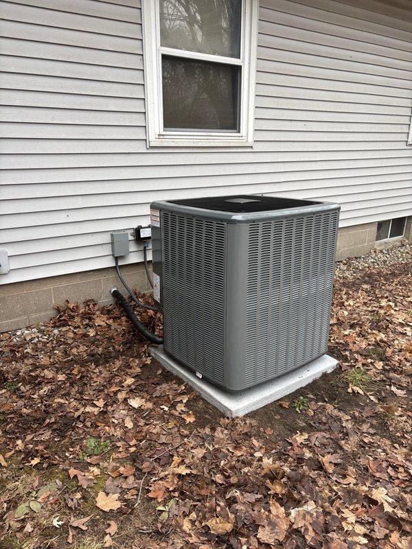 Completed full HVAC system replacement including outdoor condensing unit and indoor air handler. New gray condensing unit installed on existing concrete pad with proper refrigerant line connections, electrical disconnect, and conduit routing to exterior wall. Indoor air handler installed in utility area with digital thermostat control mounted on adjacent wall, filter access panel positioned at top of unit for maintenance accessibility. Both units commissioned and operational.