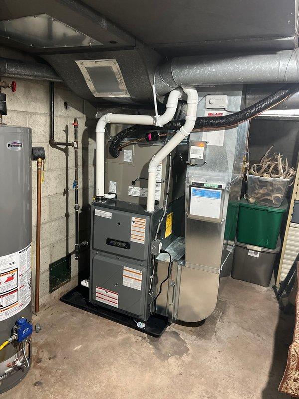 Completed full HVAC system installation including new furnace and air conditioning unit in residential basement mechanical room. Installed dark gray furnace with white PVC venting, connected to silver metallic evaporator coil and black flexible ductwork routed along ceiling joists. Mounted outdoor condensing unit on concrete pad against exterior wall with proper refrigerant line connections, electrical disconnect, and secured copper line set penetration through siding.