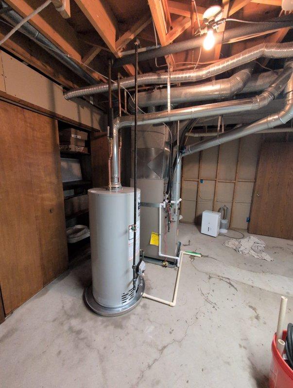 Successfully completed water heater installation in basement mechanical room. Removed existing unit and installed new white cylindrical water heater with proper venting and connections. Connected metal vent pipe extending to ceiling ductwork system, ensuring proper exhaust ventilation. Verified all plumbing connections, gas lines, and safety features are functioning correctly. Unit positioned on concrete floor alongside existing HVAC equipment with adequate clearance for maintenance access. All manufacturer specifications followed during installation, including proper TPR valve installation and drain pan placement. System tested and operational upon completion.