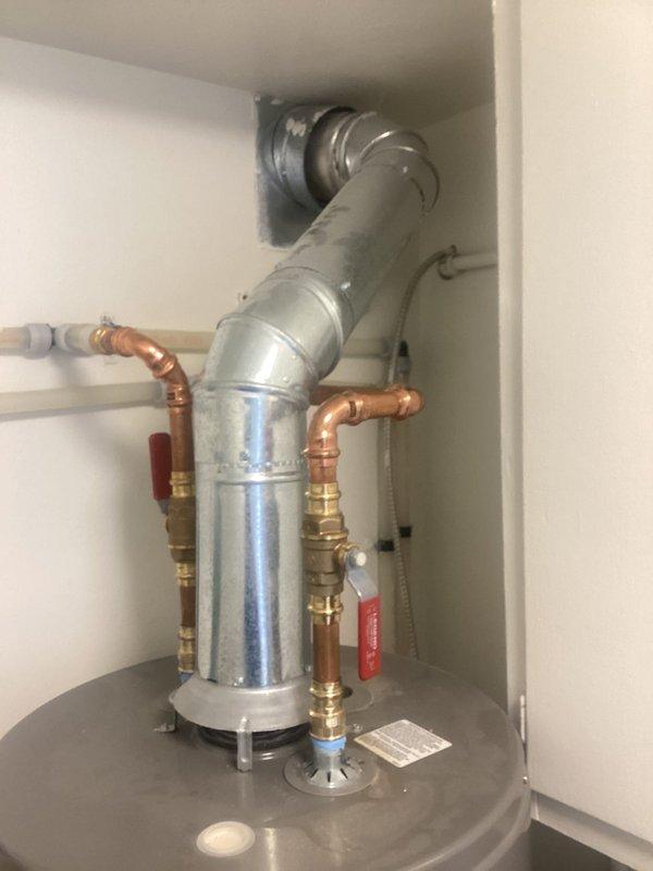 Responded to reported leak around hot water heater in 1951-built residence. Upon inspection, found no active water present at time of visit. Examined water heater cabinet installation including all copper piping, brass fittings, and connections. Inspected vent pipe system extending from water heater through ceiling into attic space. Accessed attic to assess condition of insulated chimney chase and vent termination point. Documented all plumbing connections, shut-off valves, and venting components with photographs for reference. Confirmed proper installation of galvanized vent duct with insulation wrap and structural supports. No visible signs of active leaking observed during inspection period.