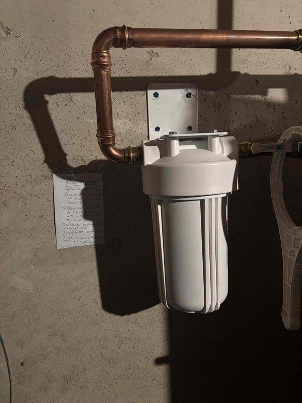 Returned to property to correct the mounting installation of a wall-mounted water filter housing. The filter's mounting bracket had been improperly secured to the overhead support structure, requiring adjustment of the chrome mounting bolts and repositioning of the white metal mounting plate against the ceiling beam. Verified proper alignment of the filter housing with the copper supply piping and ensured all mounting hardware was tightened to manufacturer specifications for secure, long-term installation.