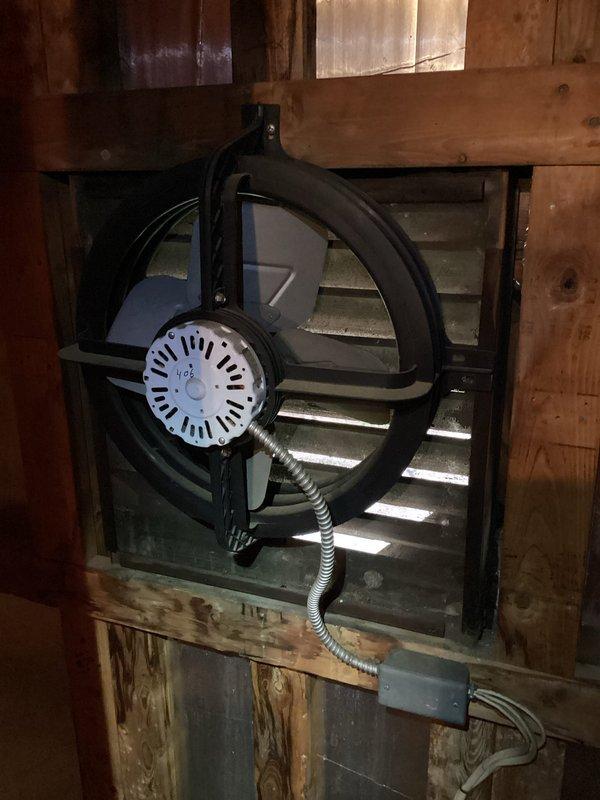 Responded to water leak complaint regarding attic fan over garage. Upon inspection, found condensation dripping from attic ventilation fan unit. System was shut off at time of arrival to prevent further water intrusion. Accessed attic space and inspected fan assembly, flexible metallic ductwork, and surrounding insulation for moisture damage. Documented conditions with photos showing the circular fan unit with central motor housing mounted in wooden frame structure and silver spiral-ribbed HVAC duct running through insulated attic space. Pink fiberglass insulation present throughout area with metal access panel visible. Confirmed condensation issue related to attic fan operation and verified system remains in off position pending further evaluation or repair recommendations.