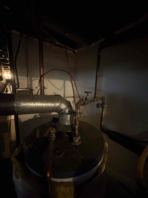 Responded to service call for water heater not producing hot water. Upon arrival, inspected residential water heater unit with blue exterior located in basement utility area. Evaluated all visible plumbing connections, copper piping, valves, and ductwork attached to the system. Documented existing installation configuration including metallic ductwork with elbow joint connections and overhead piping assembly showing signs of age and wear. Customer was provided with diagnostic findings and recommendations for resolution of heating issue.
