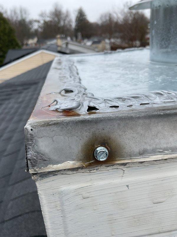 Responded to water leak complaint originating from furnace area. Upon inspection, discovered significant condensation and moisture accumulation on rooftop furnace vent pipe and flue assembly. The galvanized vent pipe exhibited heavy condensation droplets and frost formation on exterior surface, indicating inadequate insulation or excessive temperature differential between flue gases and ambient conditions. Metal flashing and mounting hardware showed corrosion and rust staining around fastener points, suggesting prolonged moisture exposure. Documented conditions with photographs showing deteriorated wood base, compromised weatherproofing, and ice/snow accumulation on flat roof membrane surrounding vent penetration. Identified condensate drainage issue as primary source of water intrusion affecting furnace equipment below.