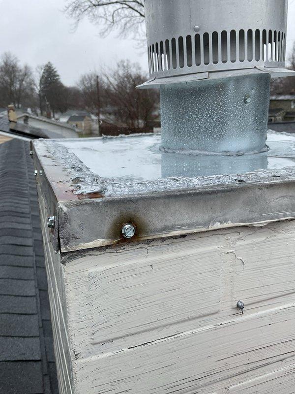 Responded to water leak complaint originating from furnace area. Upon inspection, discovered significant condensation and moisture accumulation on rooftop furnace vent pipe and flue assembly. The galvanized vent pipe exhibited heavy condensation droplets and frost formation on exterior surface, indicating inadequate insulation or excessive temperature differential between flue gases and ambient conditions. Metal flashing and mounting hardware showed corrosion and rust staining around fastener points, suggesting prolonged moisture exposure. Documented conditions with photographs showing deteriorated wood base, compromised weatherproofing, and ice/snow accumulation on flat roof membrane surrounding vent penetration. Identified condensate drainage issue as primary source of water intrusion affecting furnace equipment below.